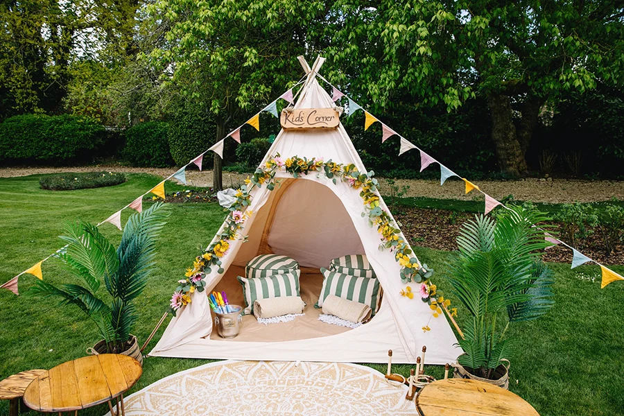 Full Kids Corner teepee with wooden “Kids Corner” sign at the top, bunting on each side, carpeted floor, wooden tables, and two plant pots, by Thistle & Rose Events.