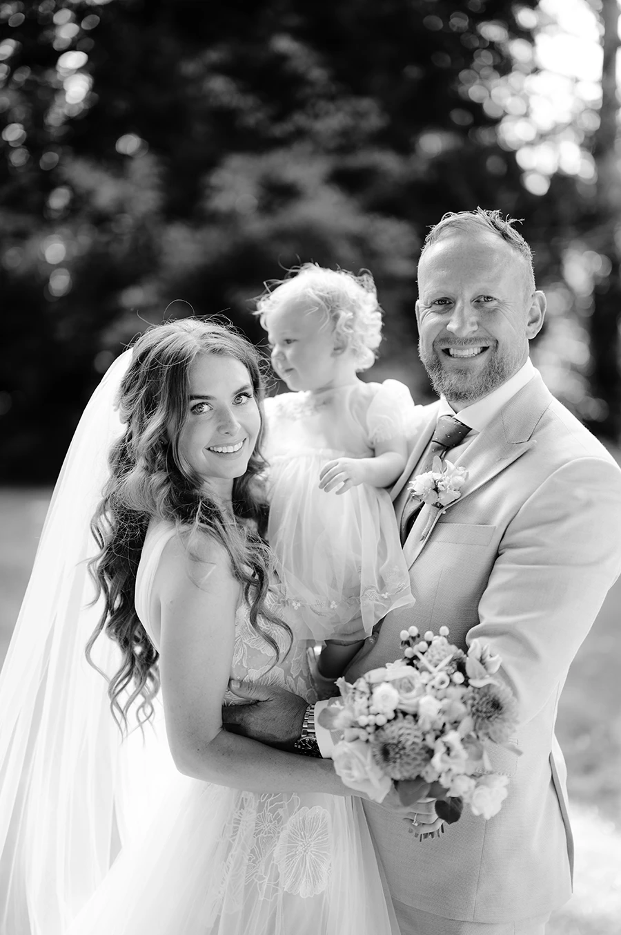 Black and white image of Rob and Sophia with their daughter on their wedding day, Sophia in wedding dress, Rob in suit, and daughter as flower girl, walking together and looking forward, by Thistle & Rose Events.